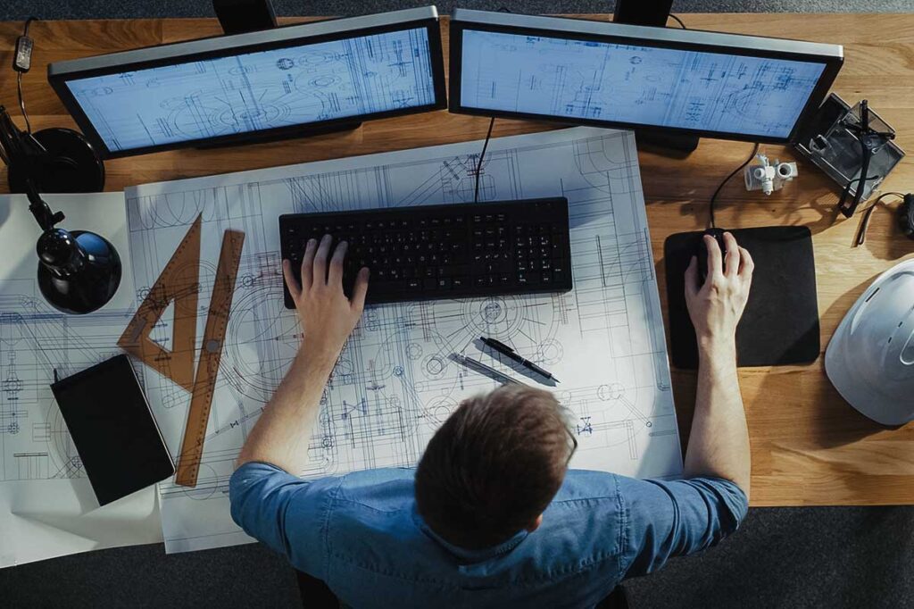 Man working at desk with 2 computer screens and a blueprint